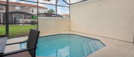 Una piscina con cerramiento de vidrio, una mesa con una silla y vistas a casas vecinas.