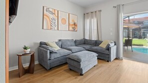 Ein modernes Wohnzimmer mit einer Eckcouch, einem kleinen Beistelltisch mit Pflanze und einem großen Fenster mit Vorhängen.