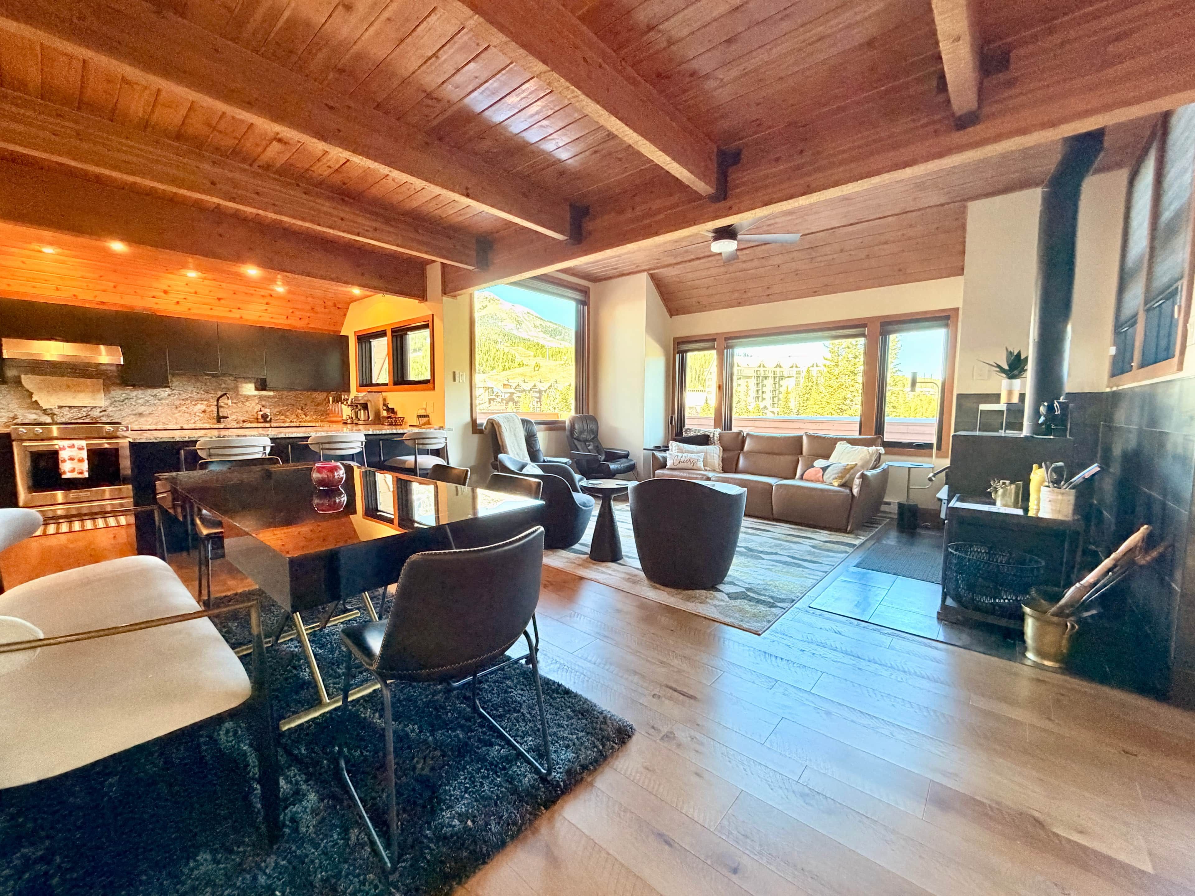 Un vaste espace de vie doté d’un plafond en bois, une cuisine équipée d’appareils électroménagers modernes et de grandes fenêtres offrant une vue sur l’extérieur.