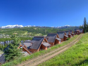 Eine Reihe von Chalets mit geneigten Dächern, vor einer Kulisse schneebedeckter Berge und blauem Himmel.