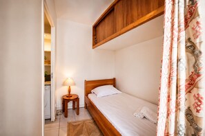 A small, single-bed room with a wooden headboard, a bedside lamp, a small round table, and a washing machine in the corner. - Studio with terrace, swimming pool and parking (Guérande)