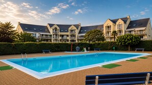 A swimming pool with a surrounding brick patio, benches, and a building with multiple balconies in the background. - Studio with terrace, swimming pool and parking (Guérande)