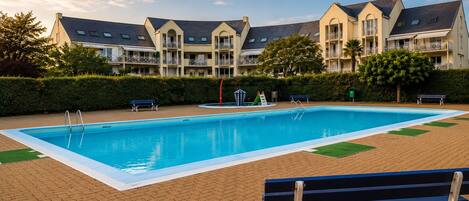 A swimming pool with a surrounding brick patio, benches, and a building with multiple balconies in the background.