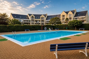 A swimming pool with a surrounding brick patio, benches, and a building with multiple balconies in the background. - Studio with terrace, swimming pool and parking (Guérande)