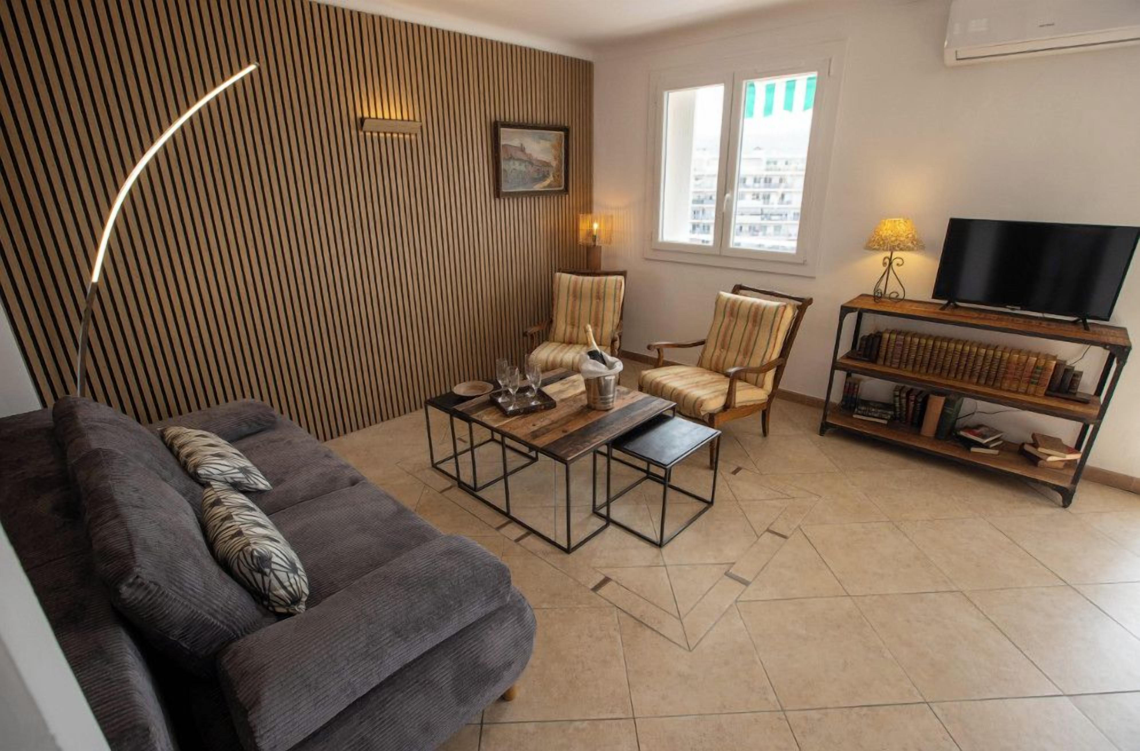 A living room with a sofa, armchairs, a coffee table, a TV stand with books, and a window with blinds.