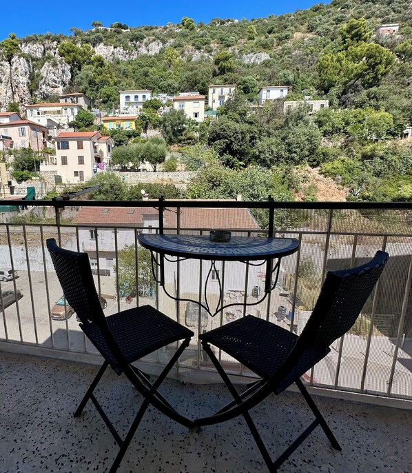 Un balcon avec deux chaises et une table, donnant sur une colline parsemée de bâtiments et d’arbres.