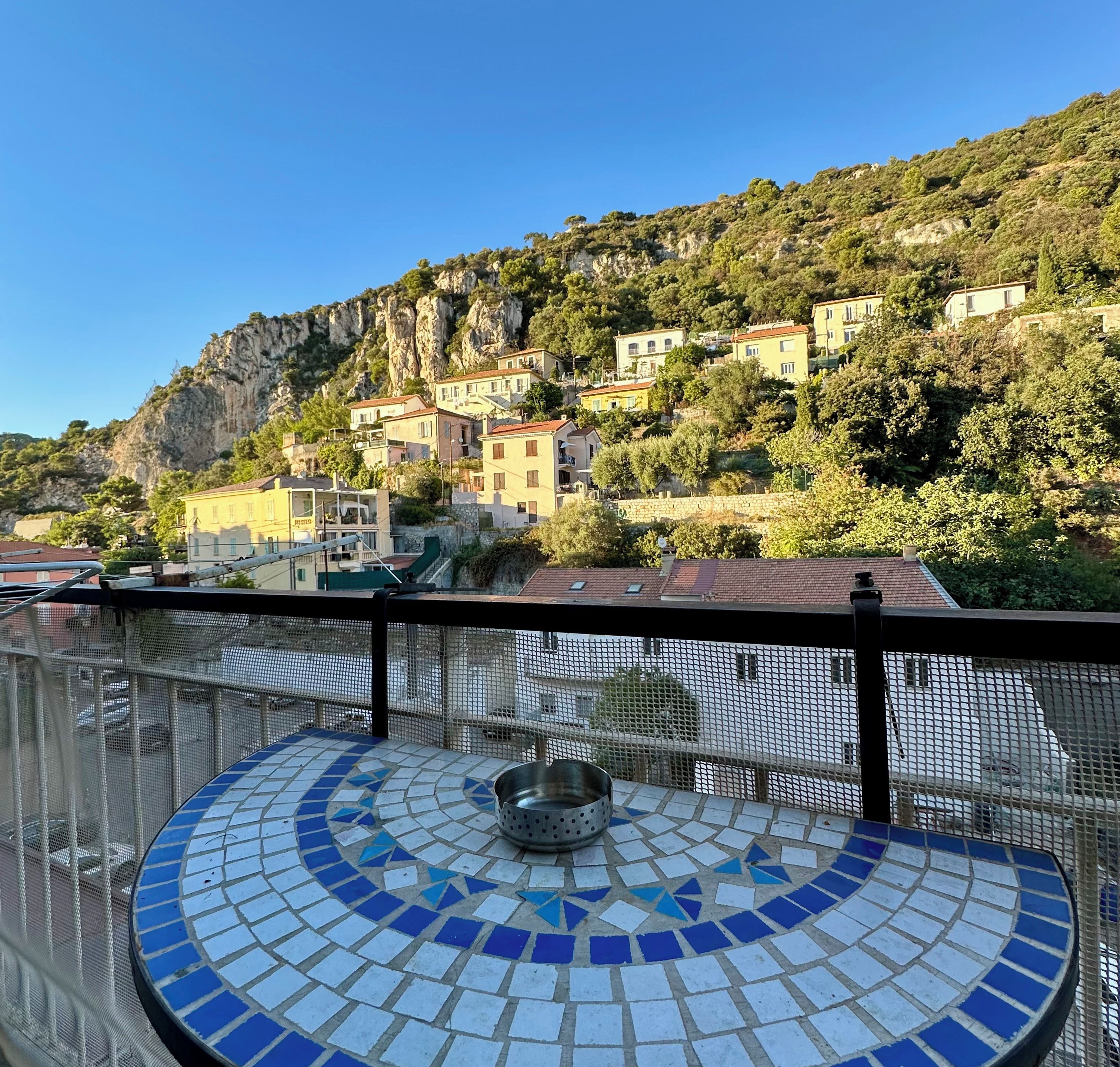 A balcony with a tiled floor, overlooking a hillside with buildings and greenery.