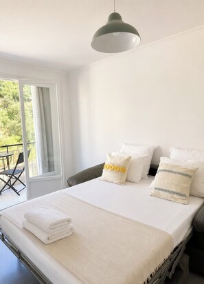 Un lit bien fait, avec du linge de lit blanc et une tête de lit grise, une suspension lumineuse et une vue sur un balcon aménagé avec du mobilier d’extérieur.