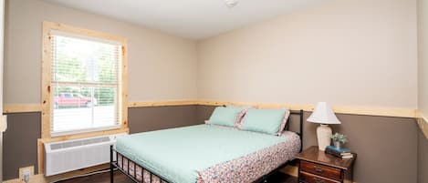 Une chambre à coucher comprenant un lit, une table de chevet, une lampe, une fenêtre avec des stores et un radiateur.