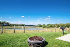 A grassy area with a metal fire pit in the foreground, a fenced pond in the background, and a building to the left. - Located in Peoria, Illinois Legacy Cabin 7 offers a luxury retreat immersed in nature, with stunning views of roaming bison and elk just outside your window. This 2-bedroom, 1-bath cabin features a comfortable living area with plush seating and a coz (Hanna City)