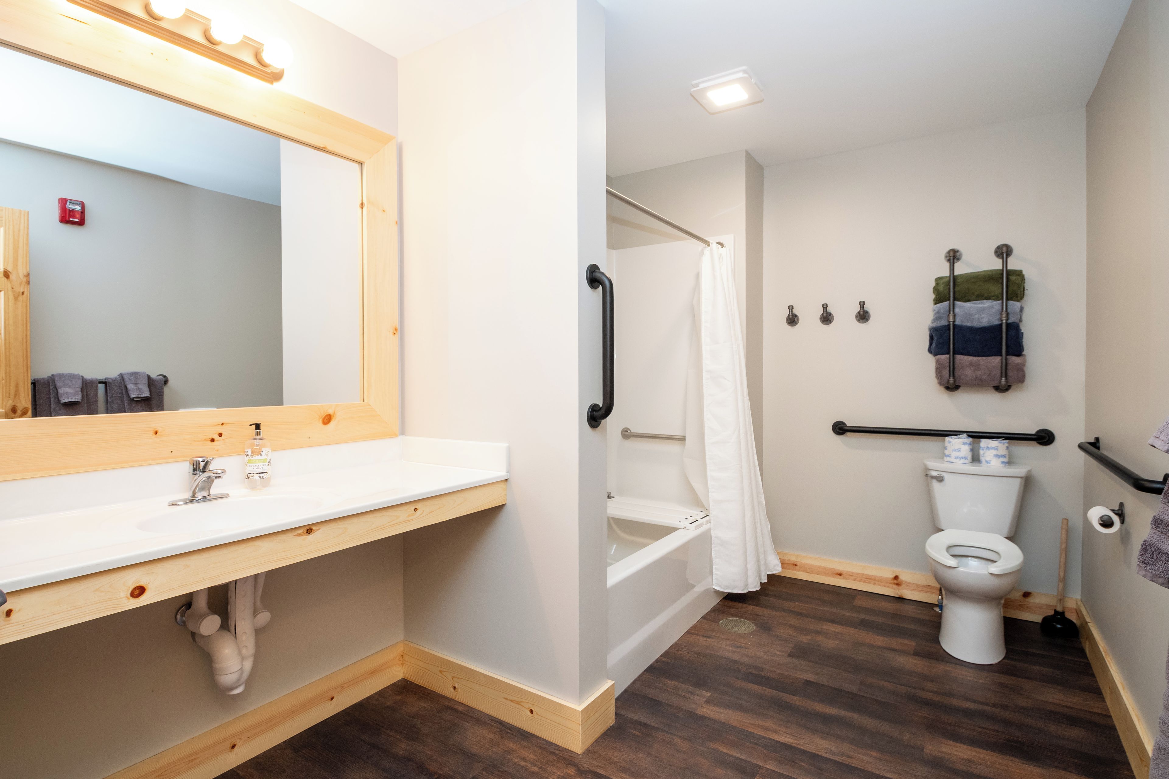 A bathroom with a wooden vanity, a white sink, a shower with a white curtain, a toilet, and a handrail.