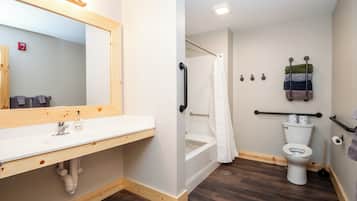 A bathroom with a wooden vanity, a white sink, a shower with a white curtain, a toilet, and a handrail.