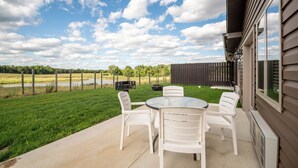 An outdoor patio with a table and chairs, a grill, and a view of a grassy field and trees. - Located in Peoria, Illinois Legacy Cabin 8 offers a luxury retreat immersed in nature, with stunning views of roaming bison and elk just outside your window. This 2-bedroom, 1-bath cabin features a comfortable living area with plush seating and a coz (Hanna City)