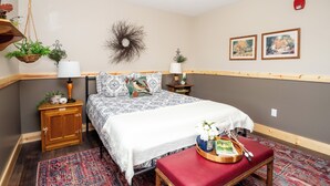 A neatly arranged bedroom with a bed, bedside tables, a hanging plant, a wall-mounted wreath, and two framed pictures. - Located in Peoria, Illinois Legacy Cabin 8 offers a luxury retreat immersed in nature, with stunning views of roaming bison and elk just outside your window. This 2-bedroom, 1-bath cabin features a comfortable living area with plush seating and a coz (Hanna City)