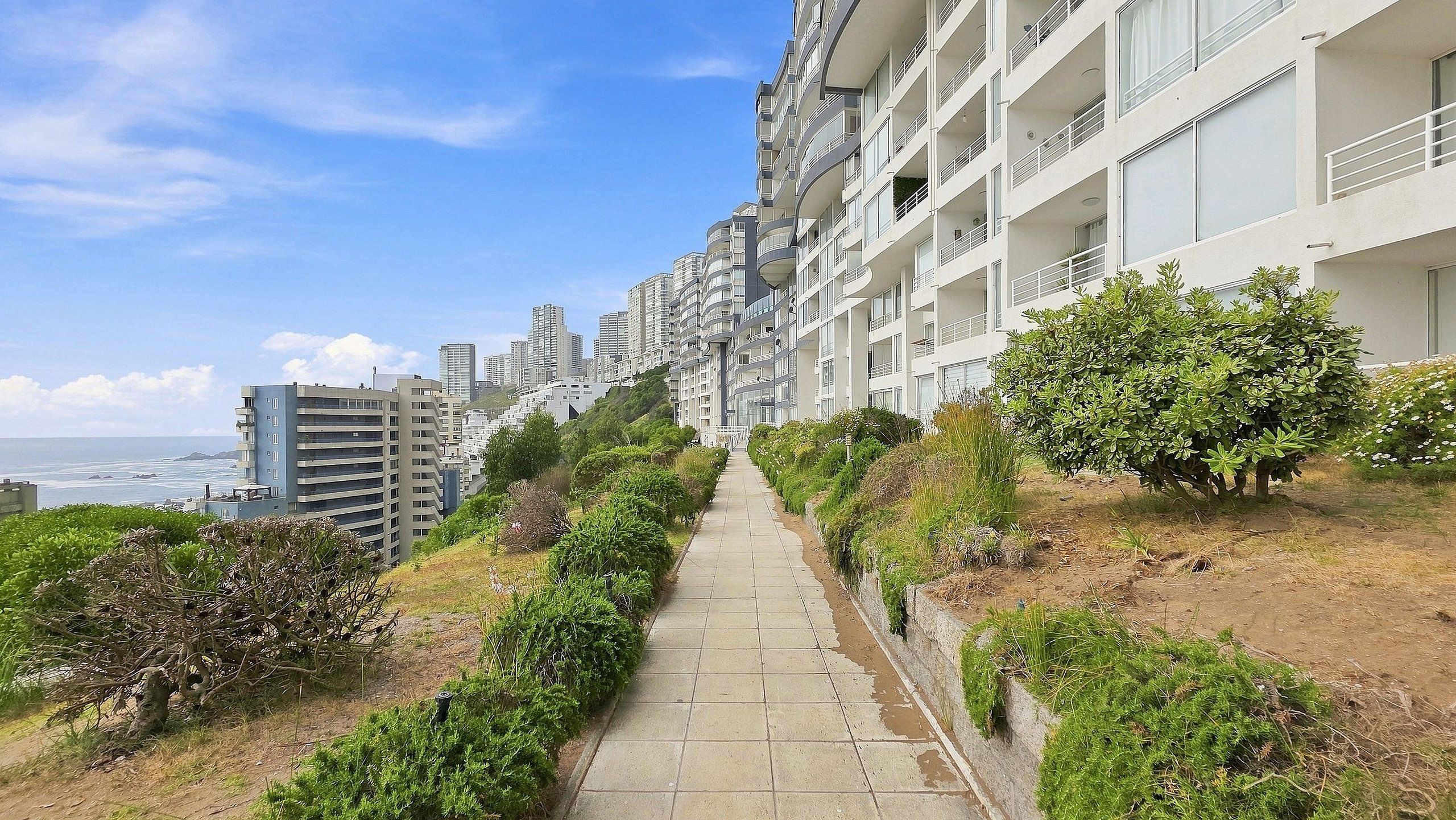 A pathway leading to a modern apartment building with balconies, surrounded by greenery and overlooking a body of water.