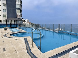 Una piscina en la azotea con barandilla de vidrio, un pequeño pabellón y vista al océano.