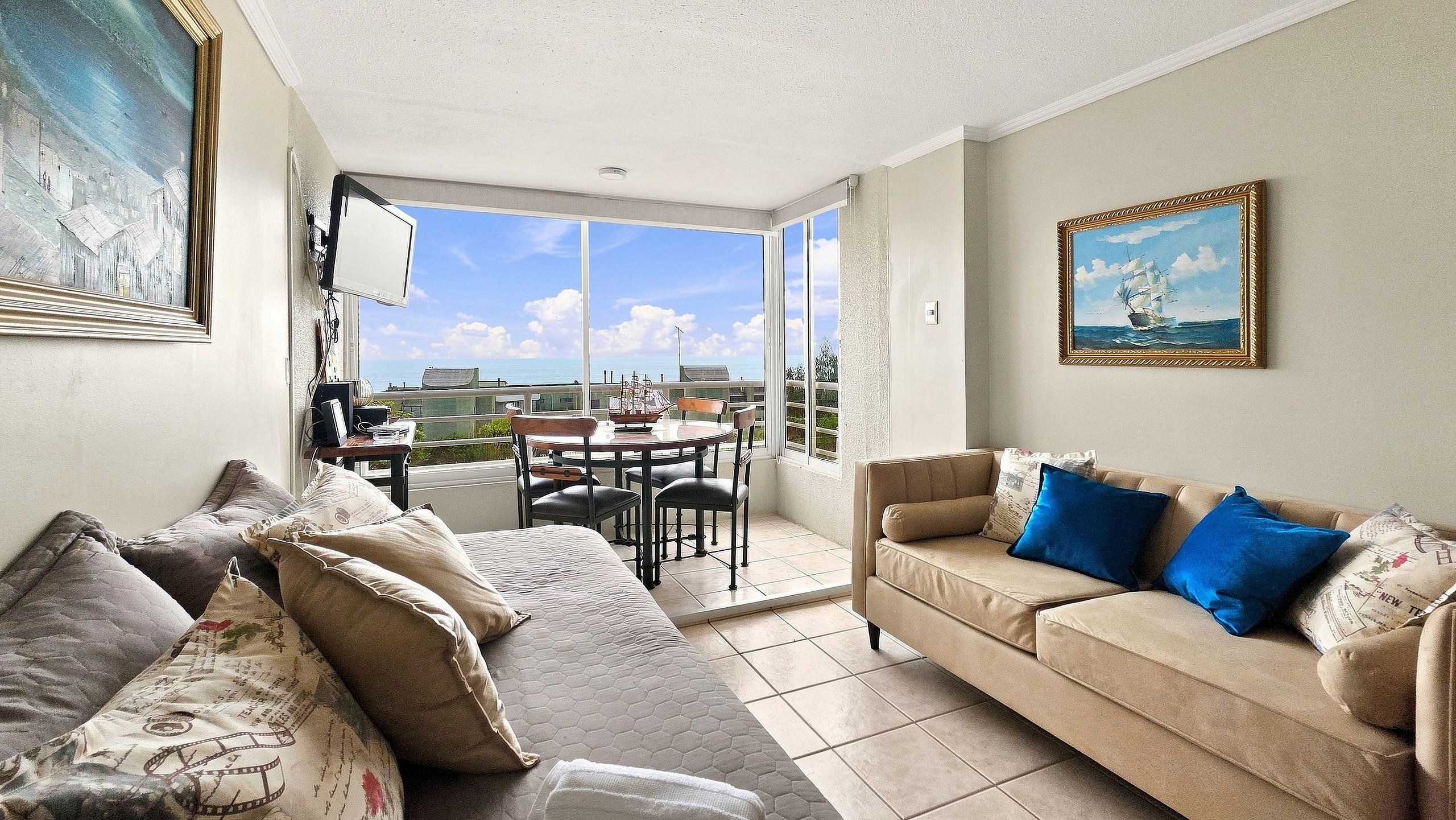 A living room with a sofa, a coffee table, and a dining table with chairs. There is a large window providing a view of the outside.