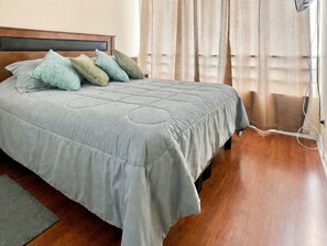 A neatly made bed with a grey quilt and multiple pillows, a wooden headboard, and a window with curtains. - Oceana Suites Euromarina II Concón (Viña del Mar)