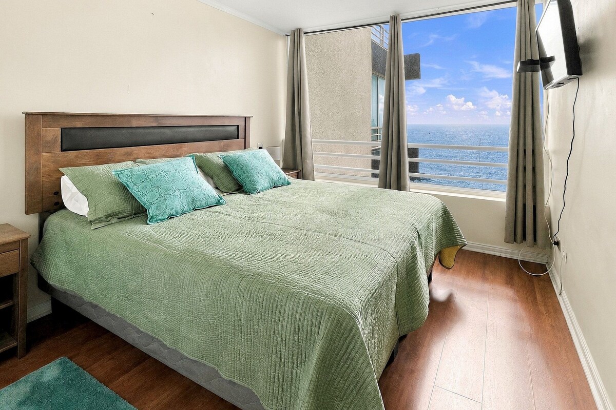 Un dormitorio con una cama grande, una mesita de noche, una ventana con vistas al mar y un televisor de pared.
