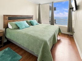Un dormitorio con una cama grande, una mesita de noche, una ventana con vistas al mar y un televisor de pared.