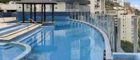 A rooftop infinity pool with a glass balustrade, a small gazebo, and a view of surrounding high-rise buildings.