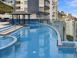 Una piscina infinita en la azotea con barandilla de cristal, un pequeño pabellón y vistas de edificios altos circundantes.