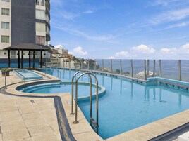 Una piscina con barrera de vidrio, zona de estar cubierta y vista al mar.
