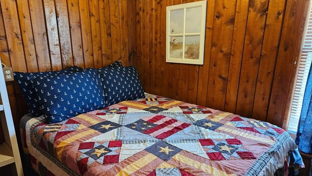 Habitación con paneles de madera, una cama con colcha de colores y almohadones azul marino con estampados de anclas.