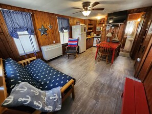 Een knus woongedeelte met een bed, een klein tafeltje, een stoel, een kitchenette en een plafondventilator.