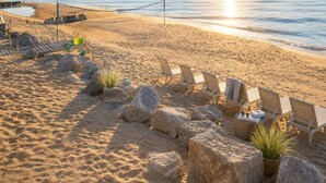 Una playa con sillas de descanso y una red de voleibol, una terraza de madera y una costa rocosa.
