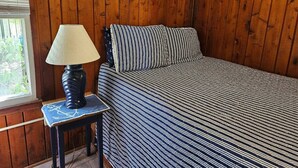 Un dormitorio con pared de paneles de madera, una cama con ropa de cama a rayas, una mesita de noche con una lámpara y una ventana con persianas.
