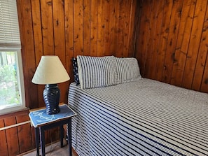 Quarto com parede de madeira, cama com roupa de cama listrada, mesa de cabeceira com abajur e janela com persianas.