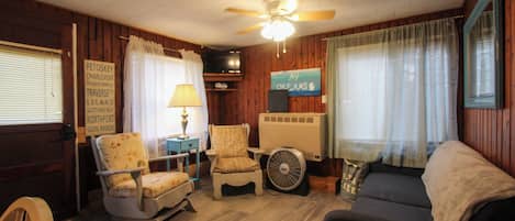 A room with a ceiling fan, two chairs, a sofa, a table with a lamp, a TV, and a sign with a list of places.