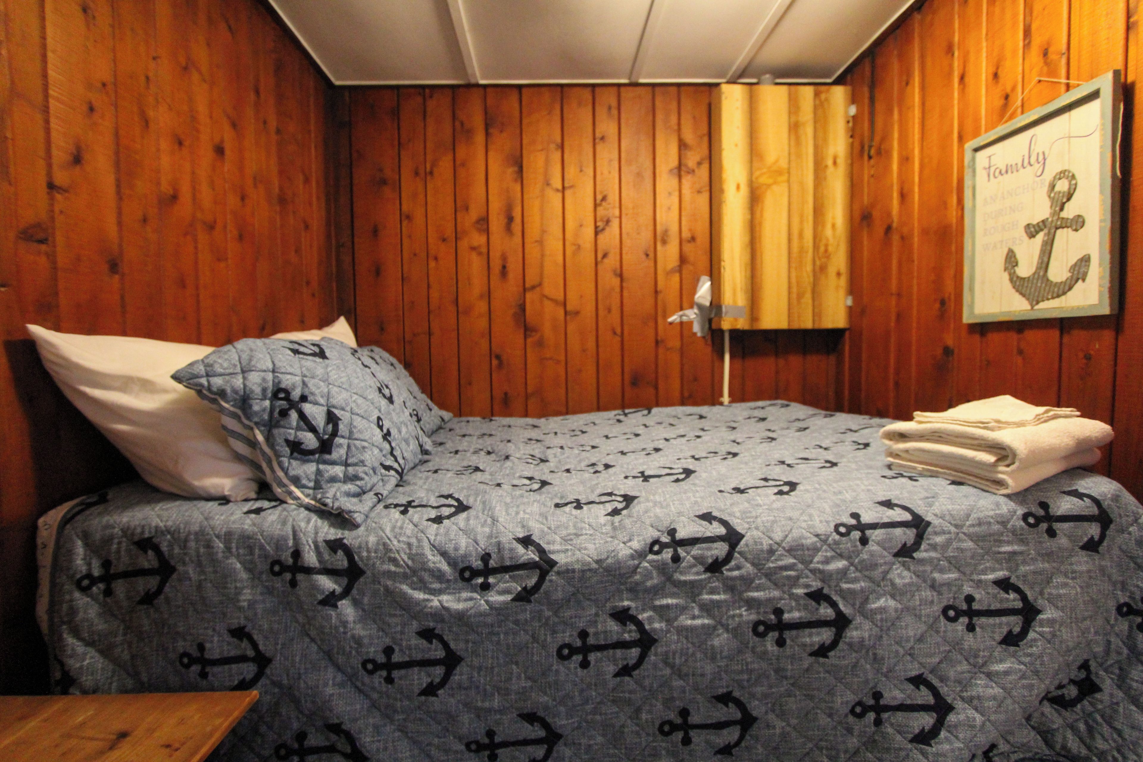 Un dormitorio con cama, almohadas, una manta, un cuadro enmarcado y una toalla doblada.