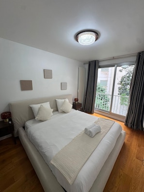 A bedroom with a large bed, a ceiling light, a window with curtains, and a view of greenery outside. - UrbanFlat 229 - Appartement Bail Mobilité Paris 16 (Paris)