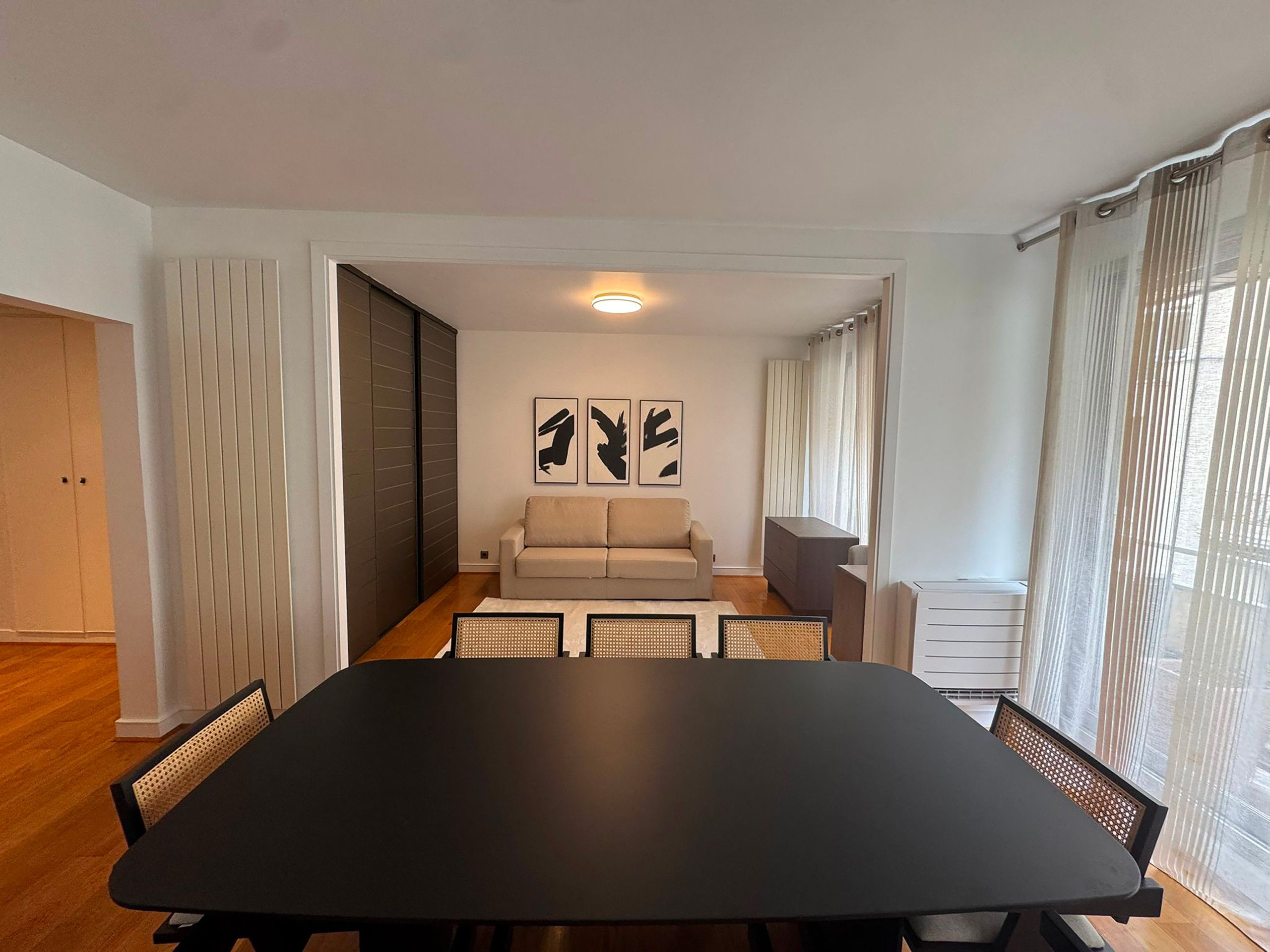 A modern living room with a large black table, two sofas, and a dining area with chairs. There are abstract wall art pieces and a window with curtains.
