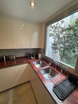 A modern kitchen with two sinks, a window with a view of trees, and a countertop with various appliances. - UrbanFlat 229 - Appartement Bail Mobilité Paris 16 (Paris)