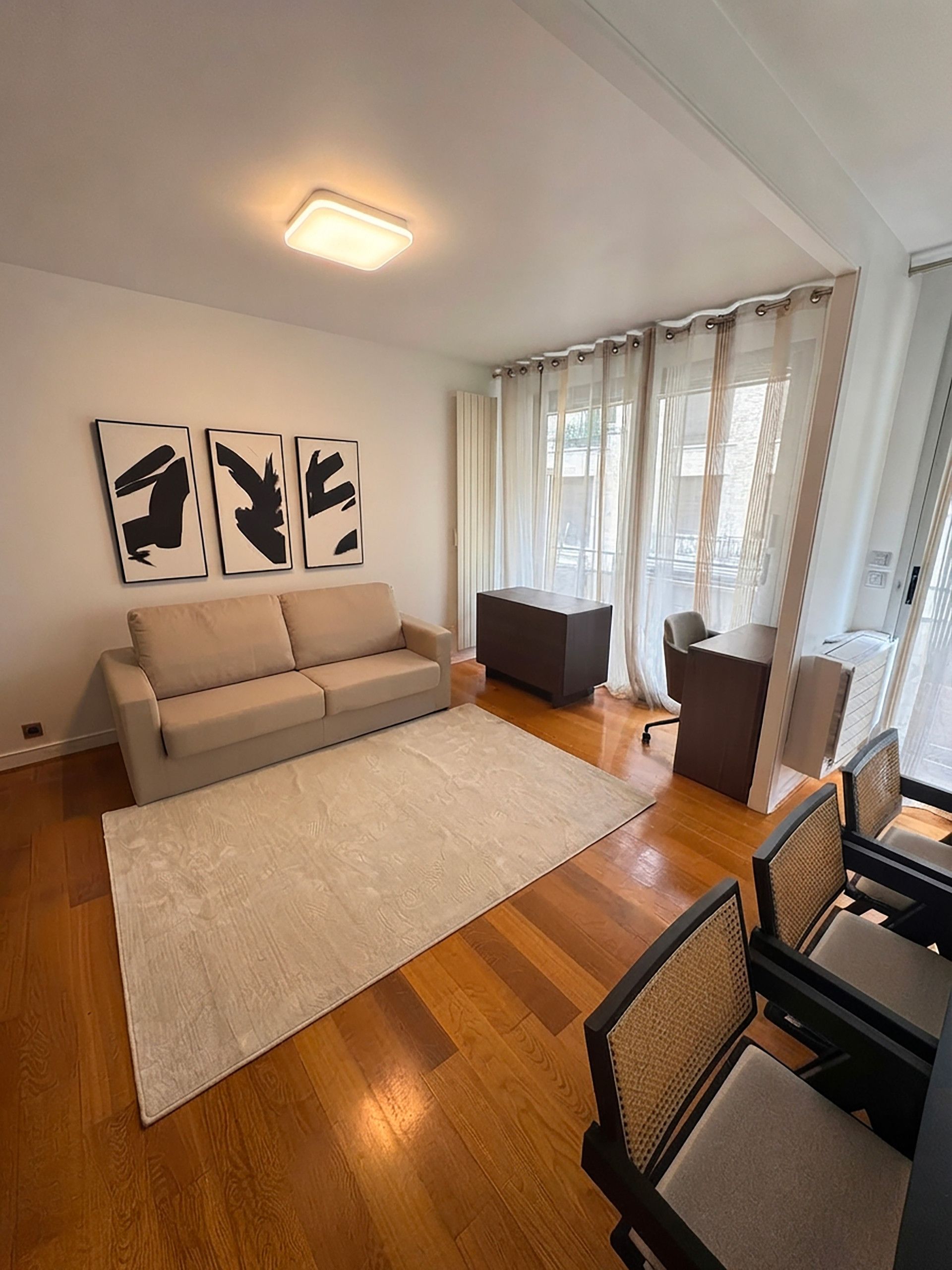 Un salon comprenant un canapé, une table basse, un tapis et un bureau avec des chaises. Trois tableaux encadrés ornent le mur.