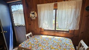 Una habitación con puerta de madera, una mesa con mantel estampado de frutas, dos sillas y una ventana con cortinas blancas.