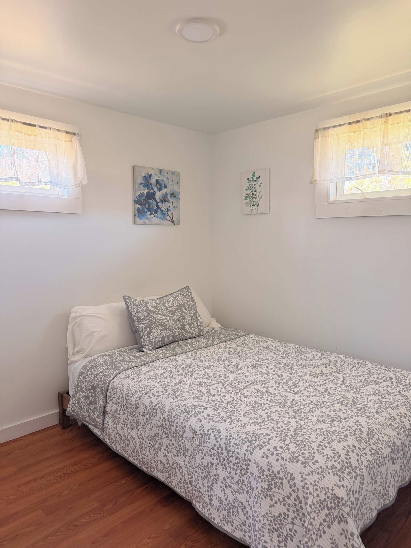A single bed with a floral-patterned bedspread in a room with white walls, a wooden floor, and two windows with sheer curtains.