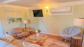 Una sala de estar con un sofá, dos sillones de mimbre, una mesa redonda, una lámpara y un televisor montado en la pared.