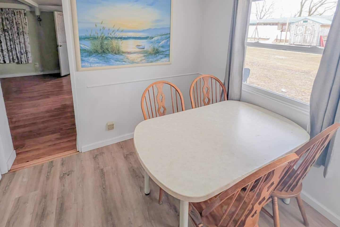 A dining area with a table and chairs, a painting on the wall, and a view through the window.