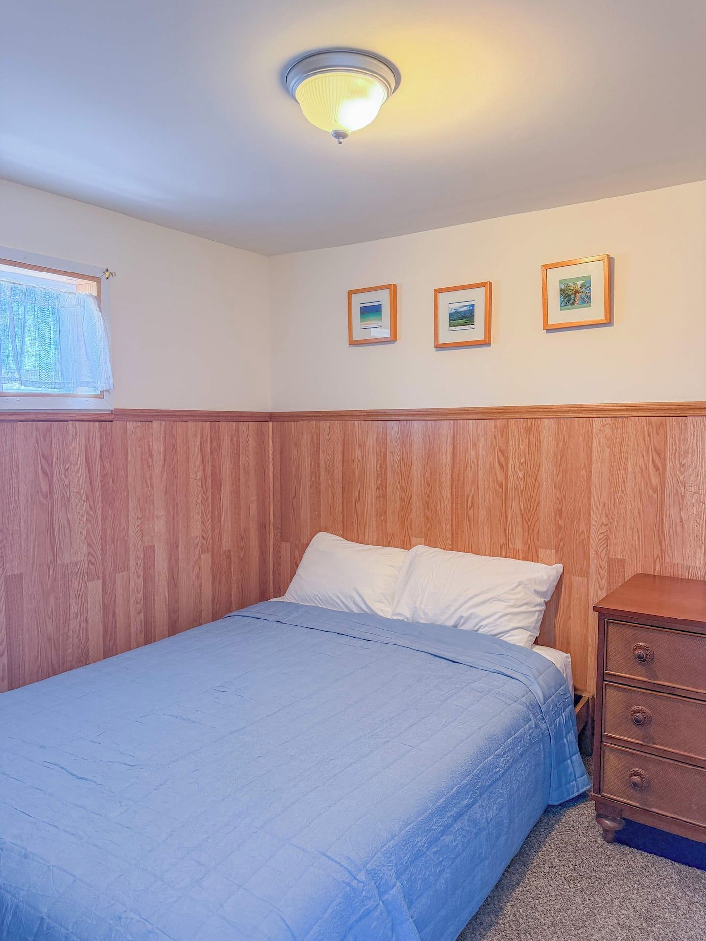 A bedroom with a wooden headboard, a bed with white bedding, a wooden dresser, and three framed pictures on the wall.