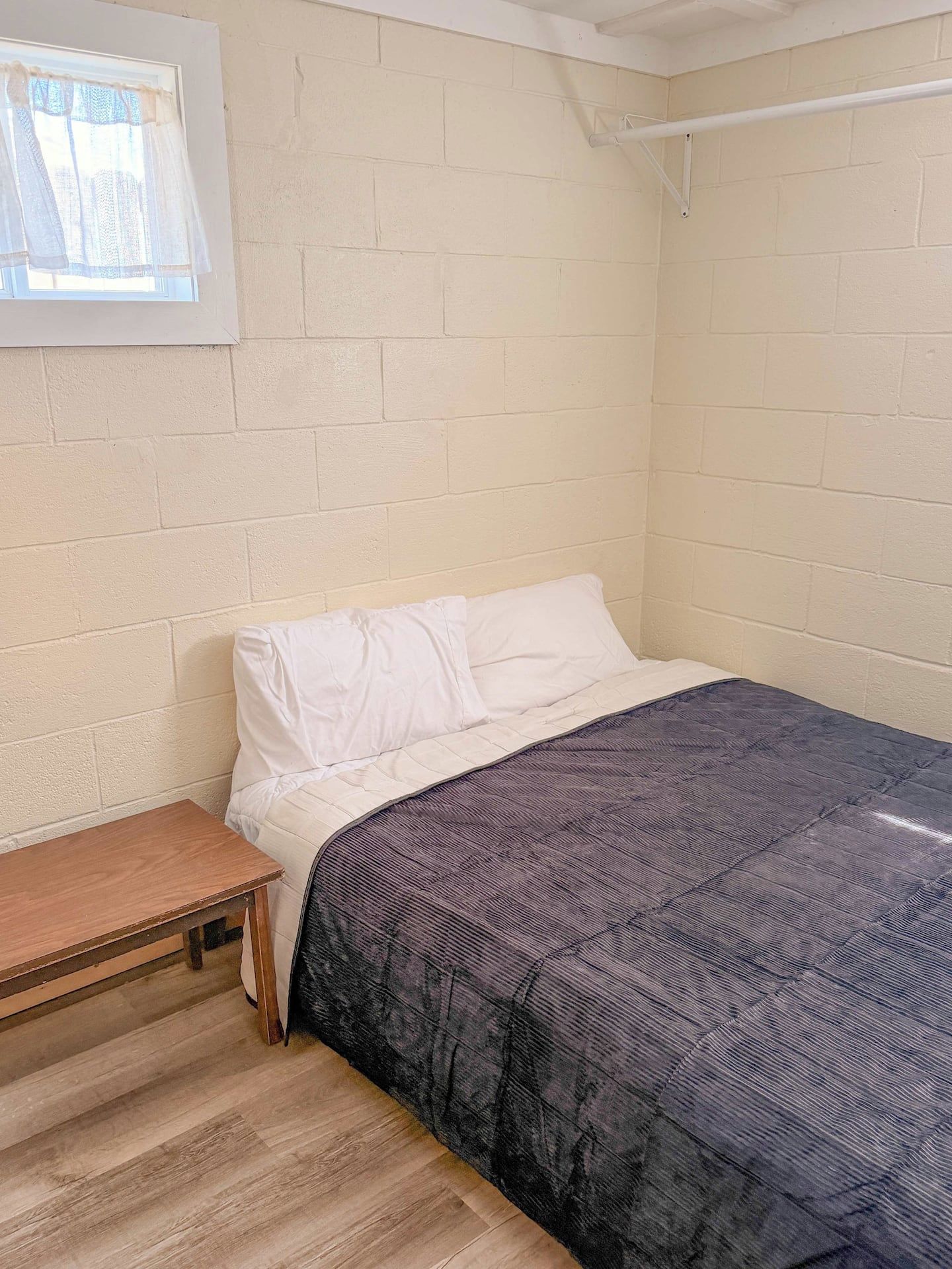 A room with a bed, a small wooden table, and a window with sheer curtains.