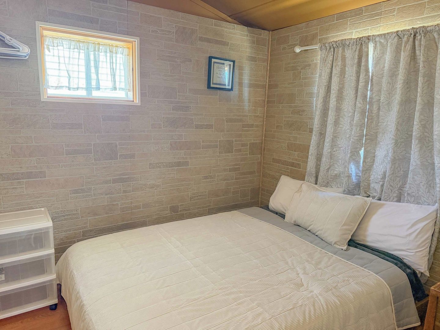 Une chambre à coucher avec un lit, une fenêtre avec des rideaux, un cadre photo fixé au mur et une tête de lit en bois.