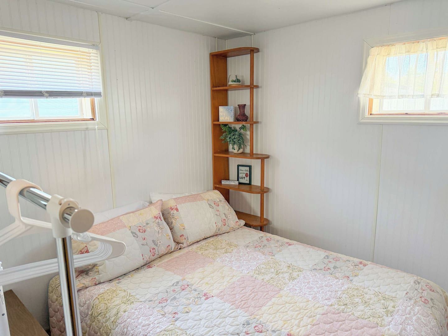 Un dormitorio con cama, mesita de noche, repisa, estante de madera con objetos decorativos y ventana con persianas.