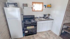 Una cocina compacta con refrigerador, estufa, horno y un microondas encima del refrigerador. Se ve una ventana con cortina, un fregadero y un estante con varios objetos.