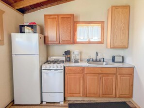 A compact kitchen with white appliances, wooden cabinets, a window with curtains, and a sink. - Charming Lake Huron Cottage | Pavilion & Firepit (Oscoda Township)