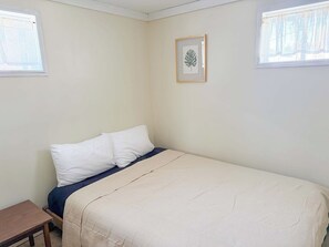 Ein Einzelbett mit weißer Matratze und einer beigen Decke, ein kleiner Nachttisch aus Holz und ein Bilderrahmen mit einem Herbstblatt an der Wand.