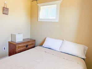 A bedroom with a bed, a nightstand, a window, and a wall-mounted sign. - Charming Lake Huron Cottage | Pavilion & Firepit (Oscoda Township)